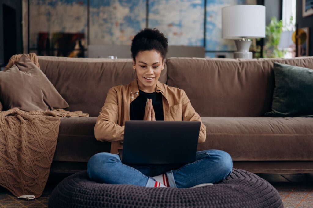 pexels photo 6894013 A smiling young woman sits cross-legged on a pouf, using her laptop in a cozy home setting.